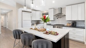 Een moderne keuken met witte kasten, een centraal eiland met een fruitschaal en kaasplateau, en hangende lampen.