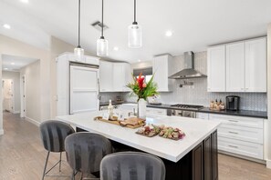Una cocina moderna con armarios blancos, una isla central con una tabla de frutas y quesos, e iluminación colgante.