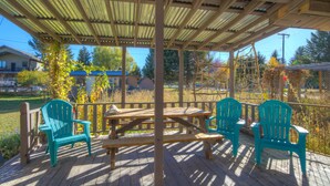 Una terraza al aire libre techada con mesas y sillas de picnic, rodeada por una cerca de madera y árboles.