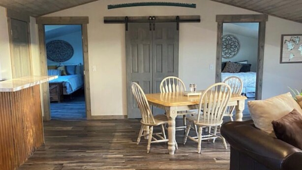 A rustic interior with a wooden floor, a dining area with a table and chairs, a ceiling fan, and a dark leather couch. - Golden Views In Every Direction! (Ennis)