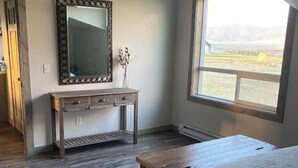 A bedroom with a large window, a wooden console table, a mirror, and a bed with a white cover. - Golden Views In Every Direction! (Ennis)
