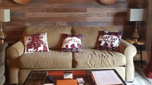 A cozy living room with a wooden wall, a mounted deer head, two lamps, a sofa with festive pillows, and a coffee table with books and a remote control.