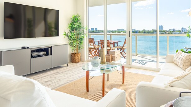 A modern living room with a large TV, a glass coffee table, and a sofa. There is a balcony with a view of the water and city skyline. - Spacious condo with beautiful water views | WIFI 100+ Mbps (Sunny Isles Beach)