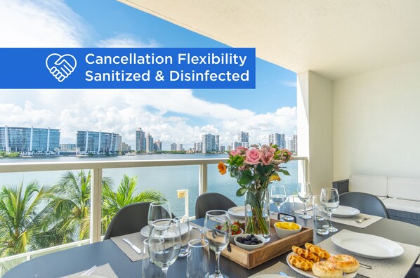 A dining area with a table set for two, a vase with flowers, and a city view. - Modern condo with amazing intracoastal views (Sunny Isles Beach)
