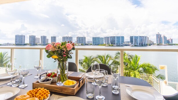 A dining table set with plates, glasses, and a waffle, overlooking a city skyline and water. - Modern condo with amazing intracoastal views (Sunny Isles Beach)
