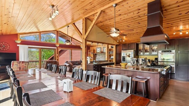 Un vaste espace de salle à manger avec une longue table et des chaises en bois, une cuisine équipée d’appareils modernes et une grande fenêtre laissant entrer la lumière naturelle.