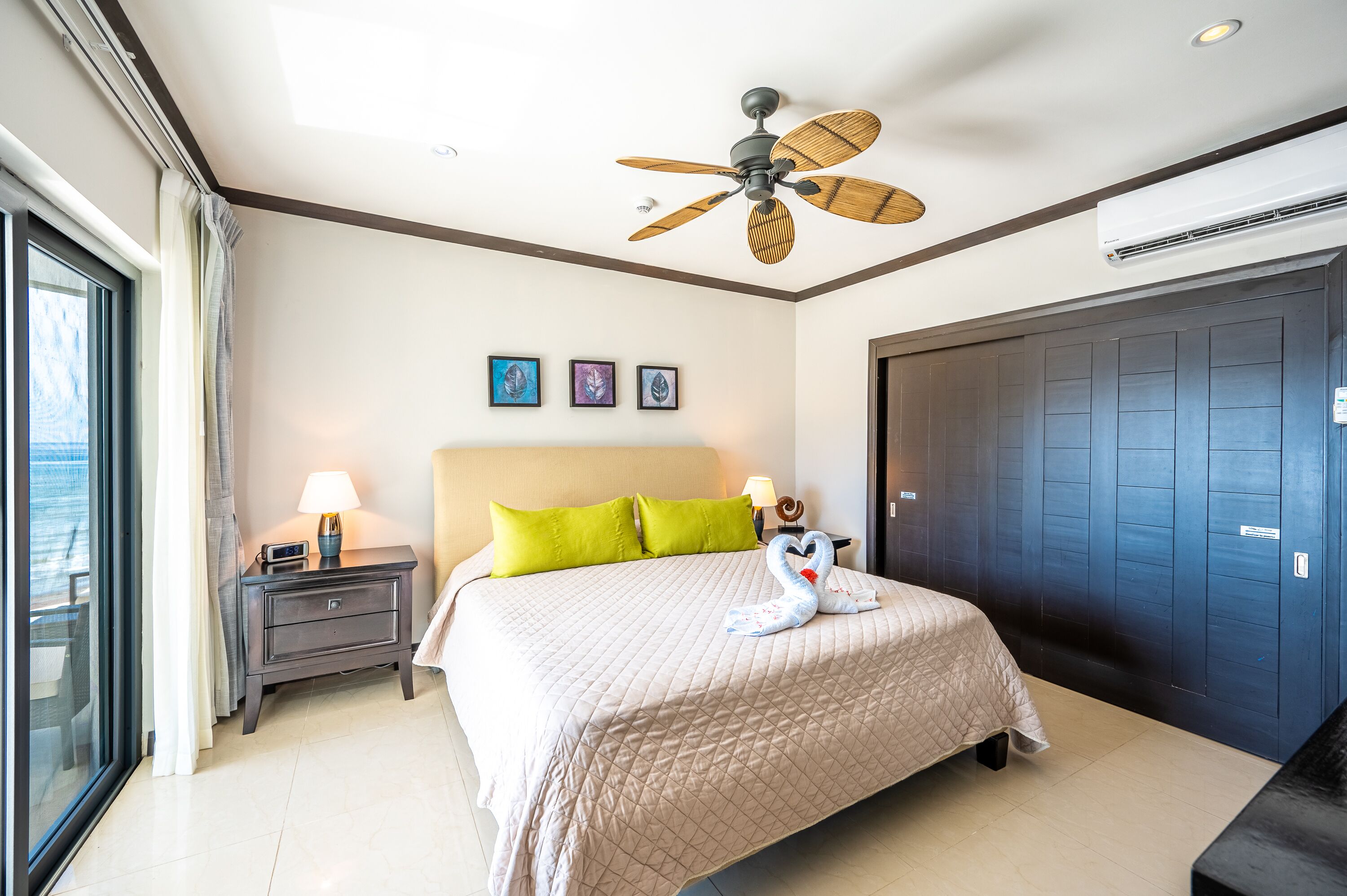 A hotel room with a bed, a nightstand, a wall-mounted air conditioner, a ceiling fan, and a large window.
