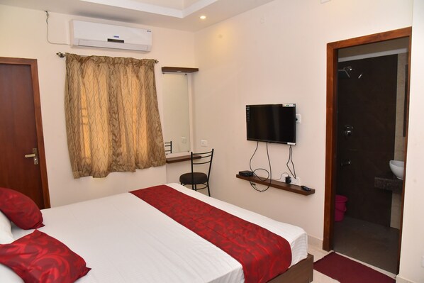 A hotel room with a bed, a TV mounted on the wall, a shower area, and a small shelf with toiletries. - Kommiya Inn (Kumbakonam)