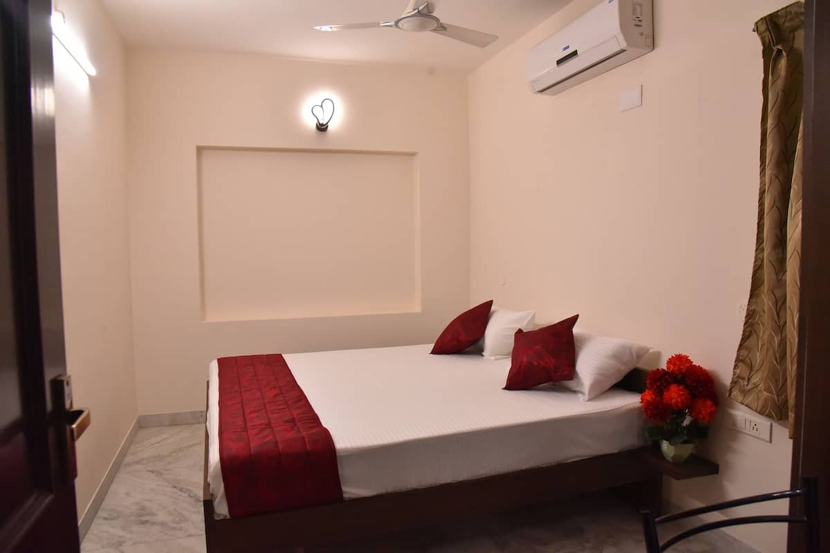 Cama bem arrumada com manta e almofadas vermelhas, um ventilador de teto, um ar condicionado e um vaso com flores vermelhas na mesa de cabeceira.