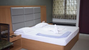 A neatly made bed with white linens and a striped towel, a wooden headboard, a wooden nightstand with a remote control, and a chair with a patterned cushion. - Hotel Vijay Krishna (Madurai)