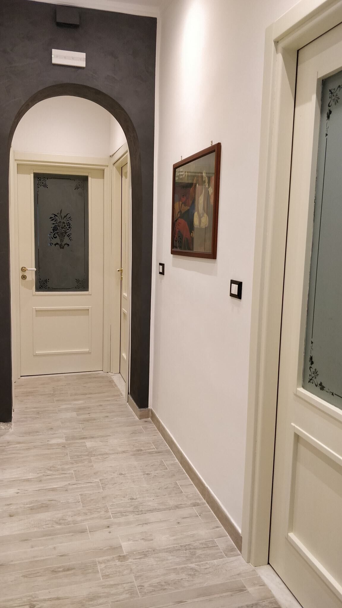 Un pasillo con suelo de madera clara, una puerta blanca con un panel de vidrio decorativo y un cuadro enmarcado en la pared.