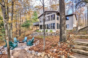 Uma cabana com lareira de pedra e cadeiras Adirondack verdes, rodeada por uma floresta e um muro de pedra.