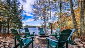 A circular fire pit with four Adirondack chairs around it, set on a stone patio with a lake and trees in the background. - Gorgeous Private Home with magnificent lake views! Great Firepit to watch the Su (Harshaw)