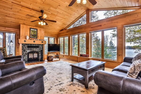 A spacious living room with a fireplace, leather sofas, a coffee table, and large windows offering a view of the outdoors. - Gorgeous Private Home with magnificent lake views! Great Firepit to watch the Su (Harshaw)