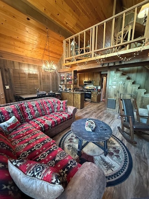 A spacious living area with a large sofa, a round coffee table, and a dining table. A kitchen is visible in the background.