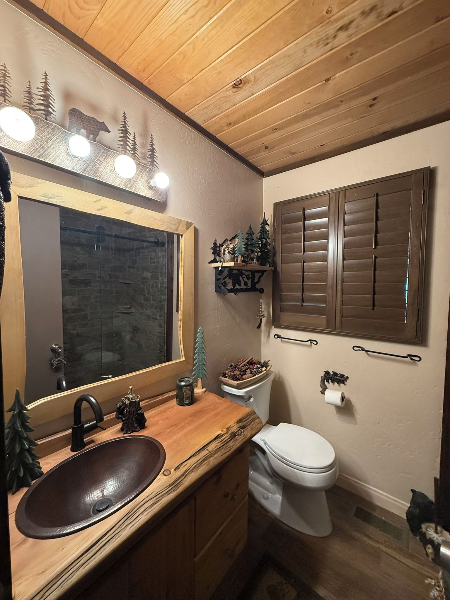 A bathroom with a wooden vanity, a mirror with lights, a toilet, and a window with shutters.