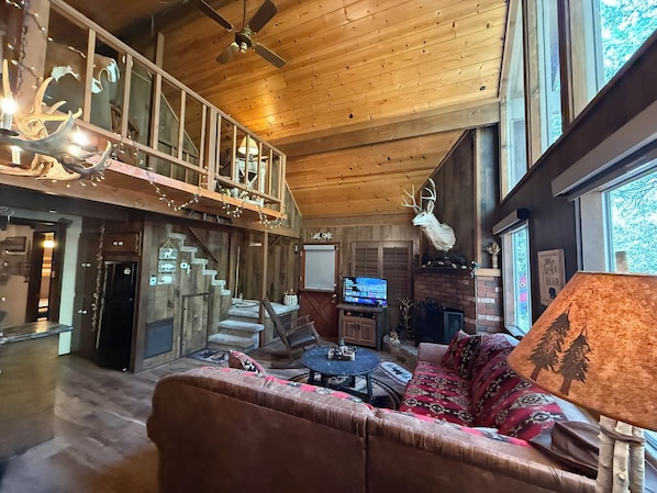A spacious living area with a high ceiling, a fireplace, a television, and a large window.