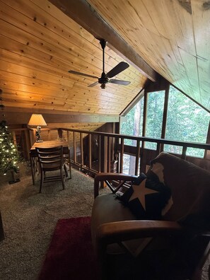 A cozy cabin interior with a wooden ceiling, a ceiling fan, a dining table with chairs, a Christmas tree, and a large window.