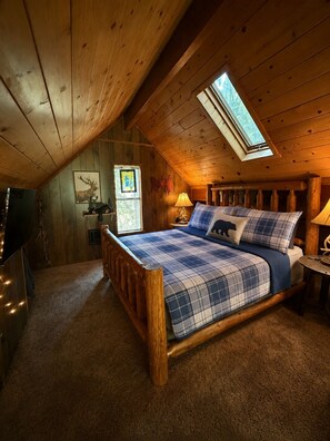 A bedroom with a wooden ceiling, a skylight, a bed with plaid bedding, and a nightstand with a lamp.