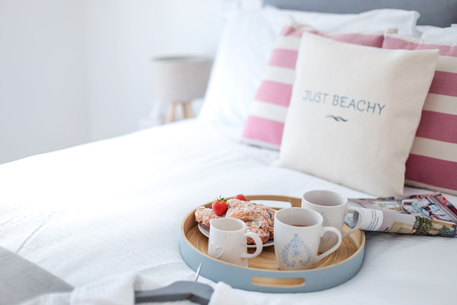 Un lit bien fait, sur lequel repose un plateau bleu contenant des pâtisseries, deux tasses blanches et un coussin rayé rose et blanc avec l’inscription « JUST BEACHY ».