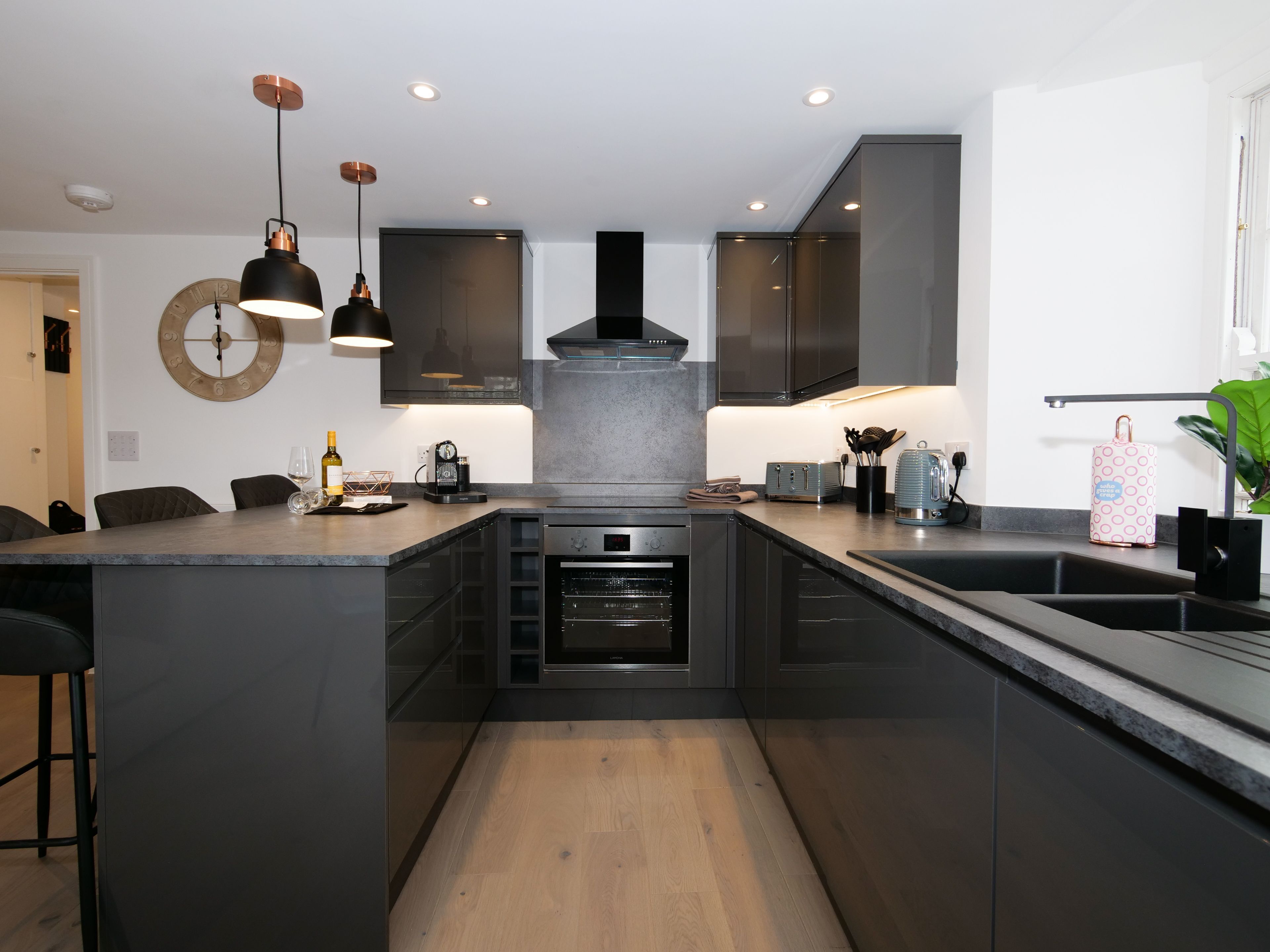 Una cocina moderna con una isla central, electrodomésticos integrados, iluminación colgante y un gran reloj en la pared.