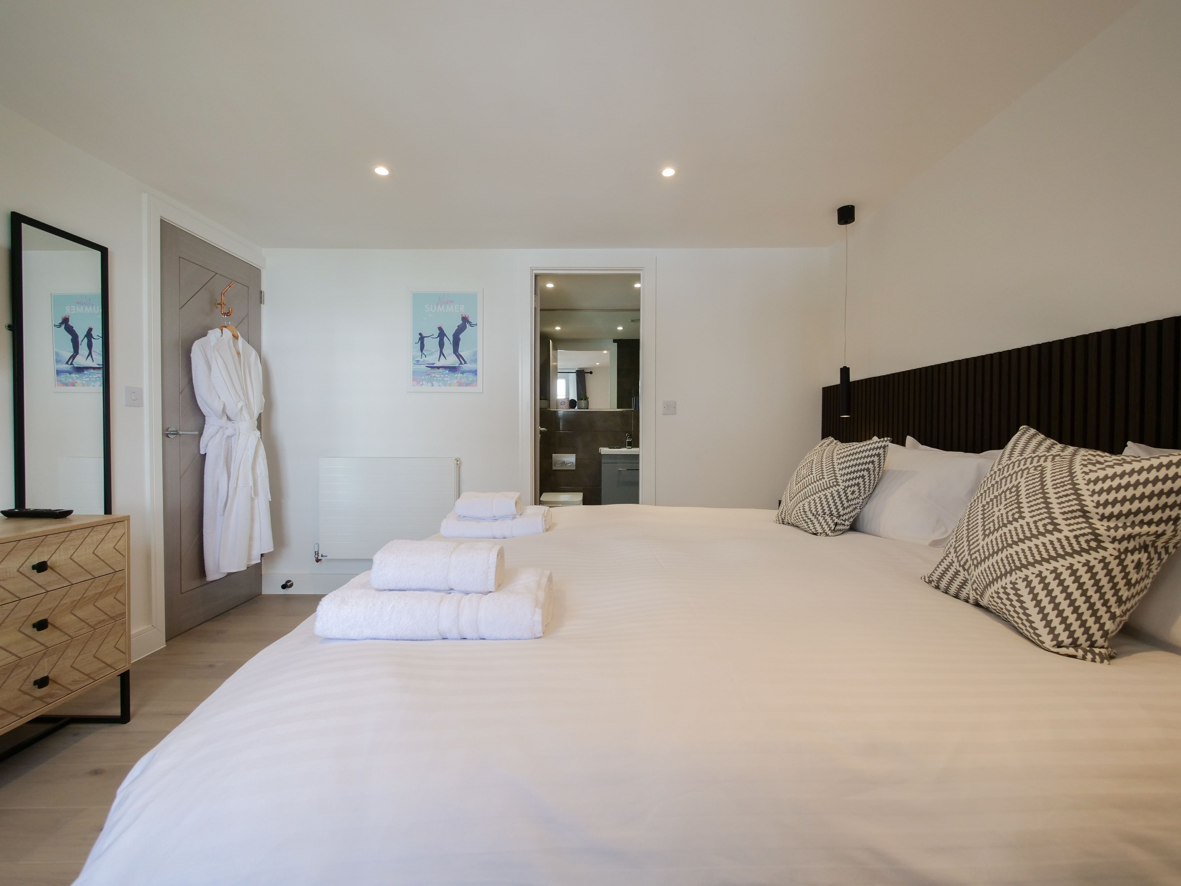 A modern bedroom with a large bed, a wooden dresser, a robe hanging on a door, and a bathroom visible through an open door.