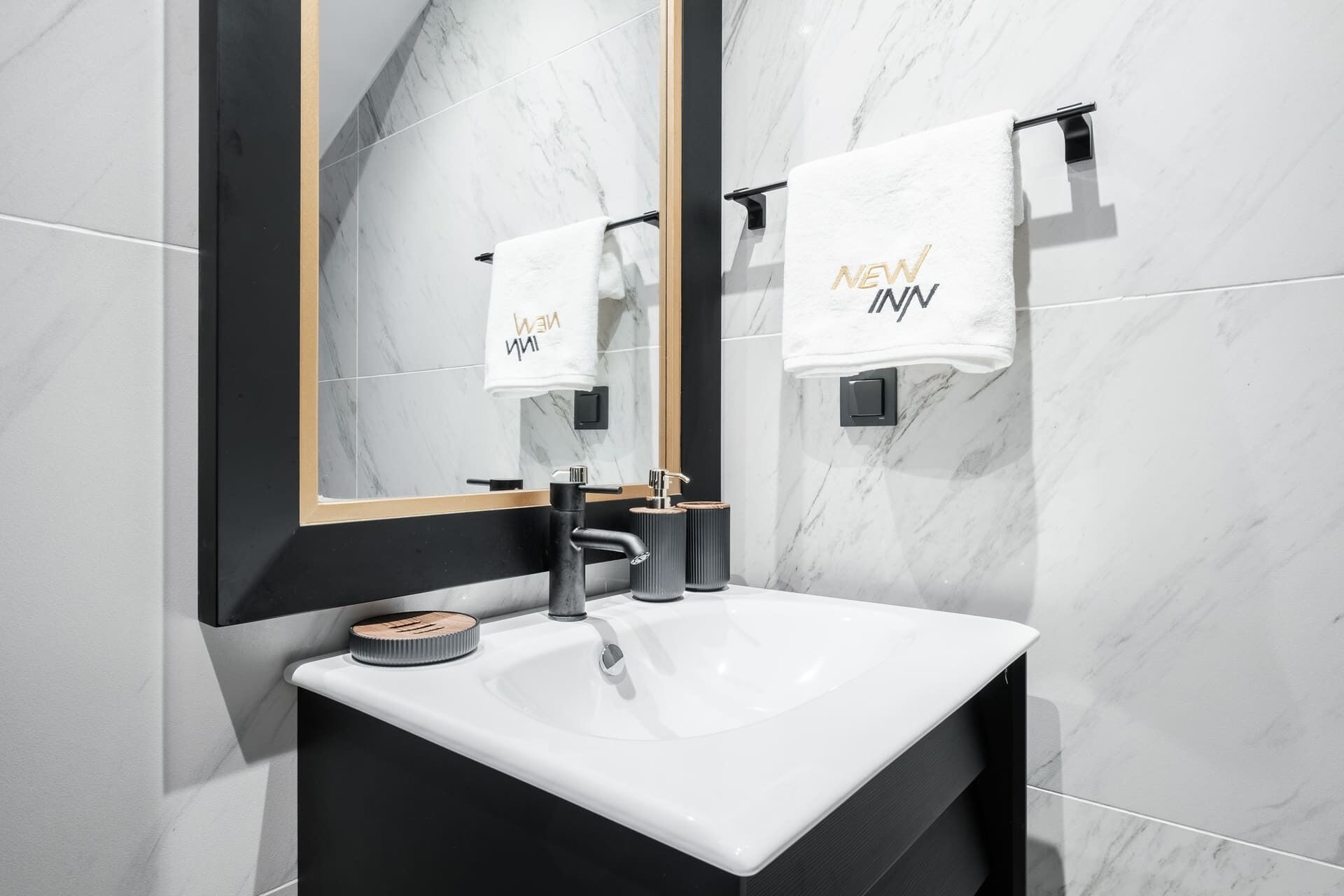 Un baño moderno con un lavamanos blanco, tocador negro y un espejo grande. Dos toallas con el texto 'NEW INN' cuelgan en la pared.