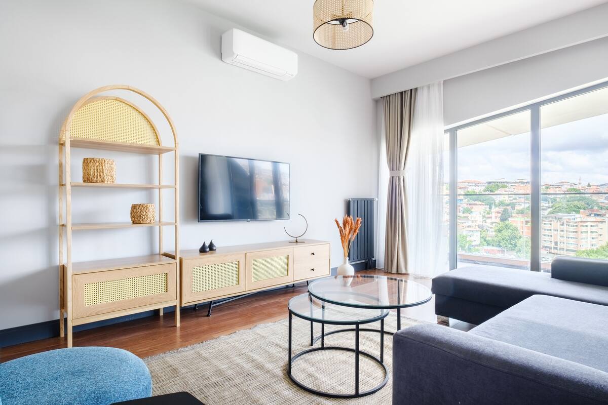 A modern living room with a large TV, a sofa, a round coffee table, and a view of the city through large windows.