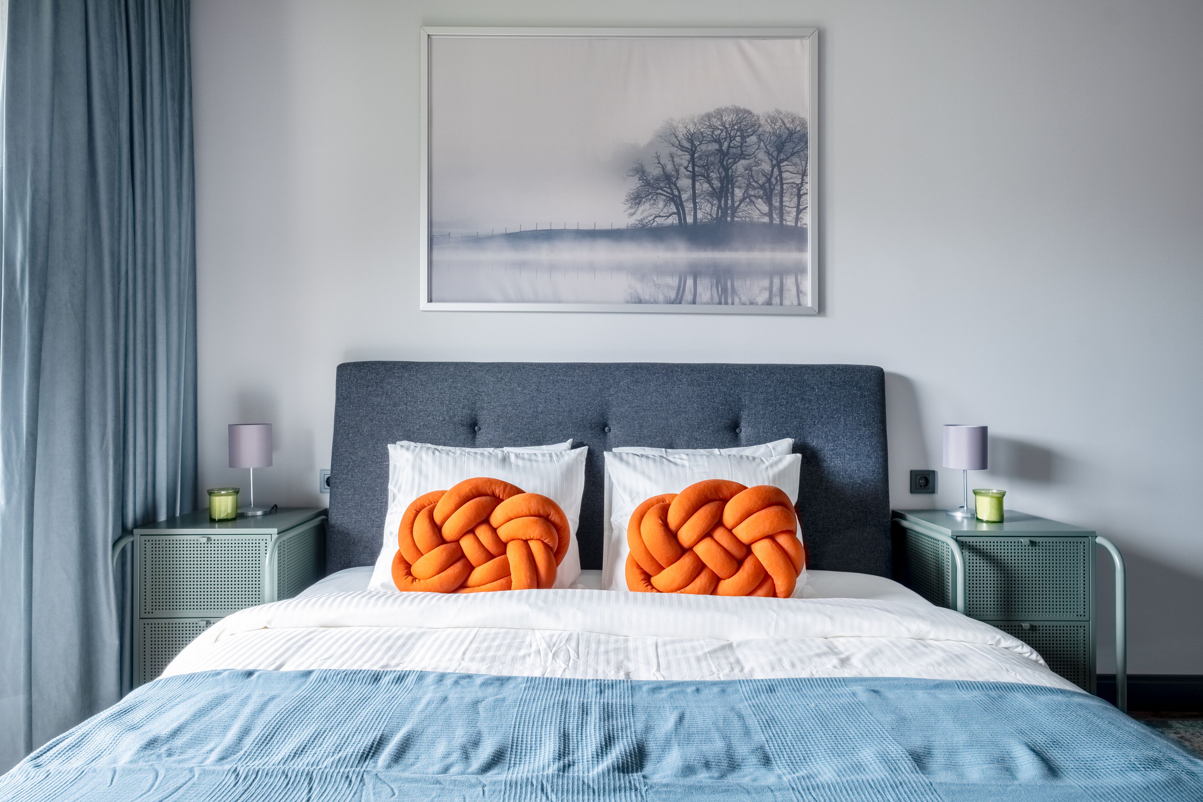 A neatly made bed with white and blue bedding, two orange braided pillows, a grey headboard, and a framed picture of a landscape on the wall.