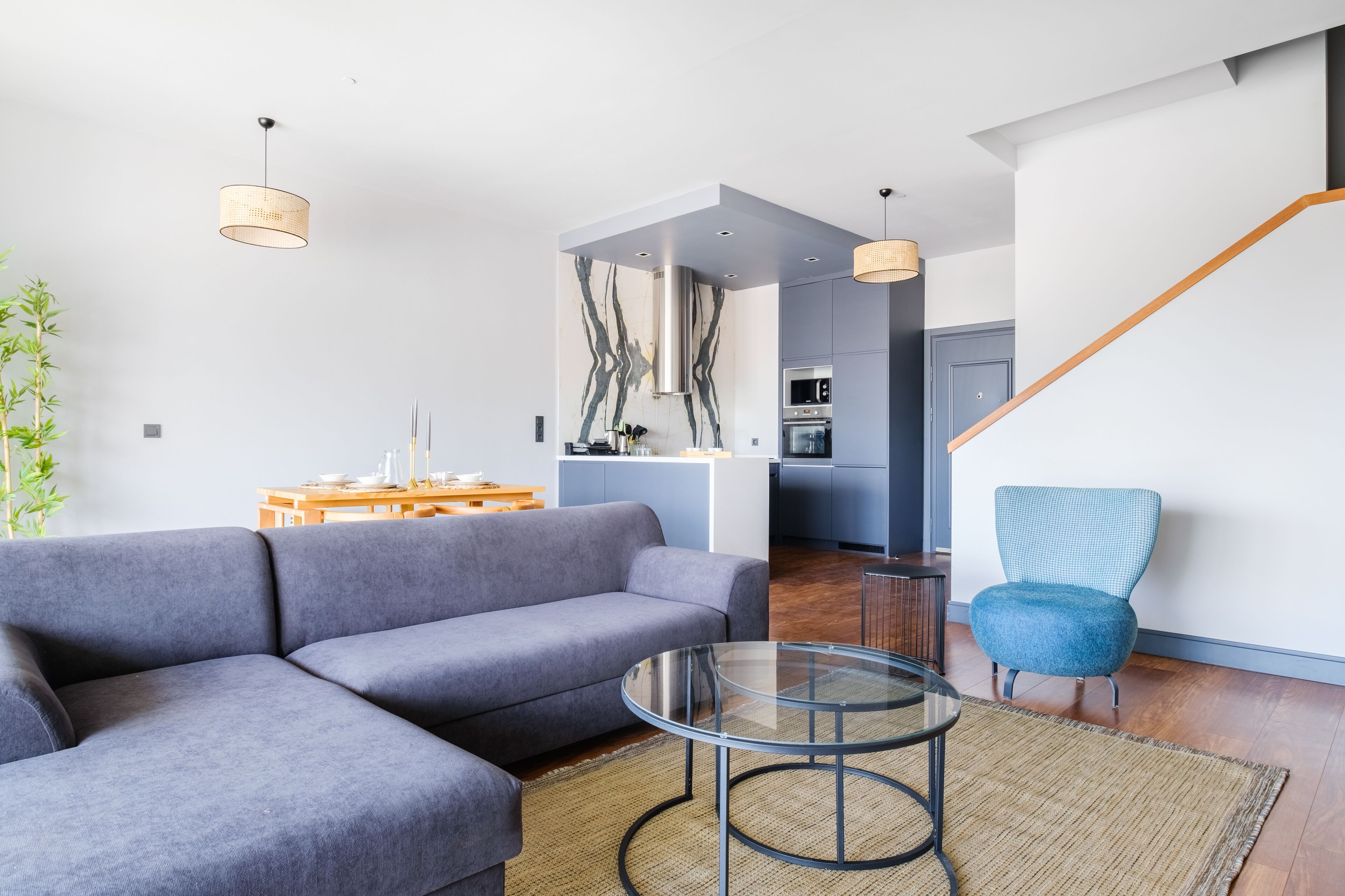 A modern living room with a grey sectional sofa, a glass coffee table, and a blue armchair. A kitchen with grey cabinets and a wooden staircase is visible in the background.