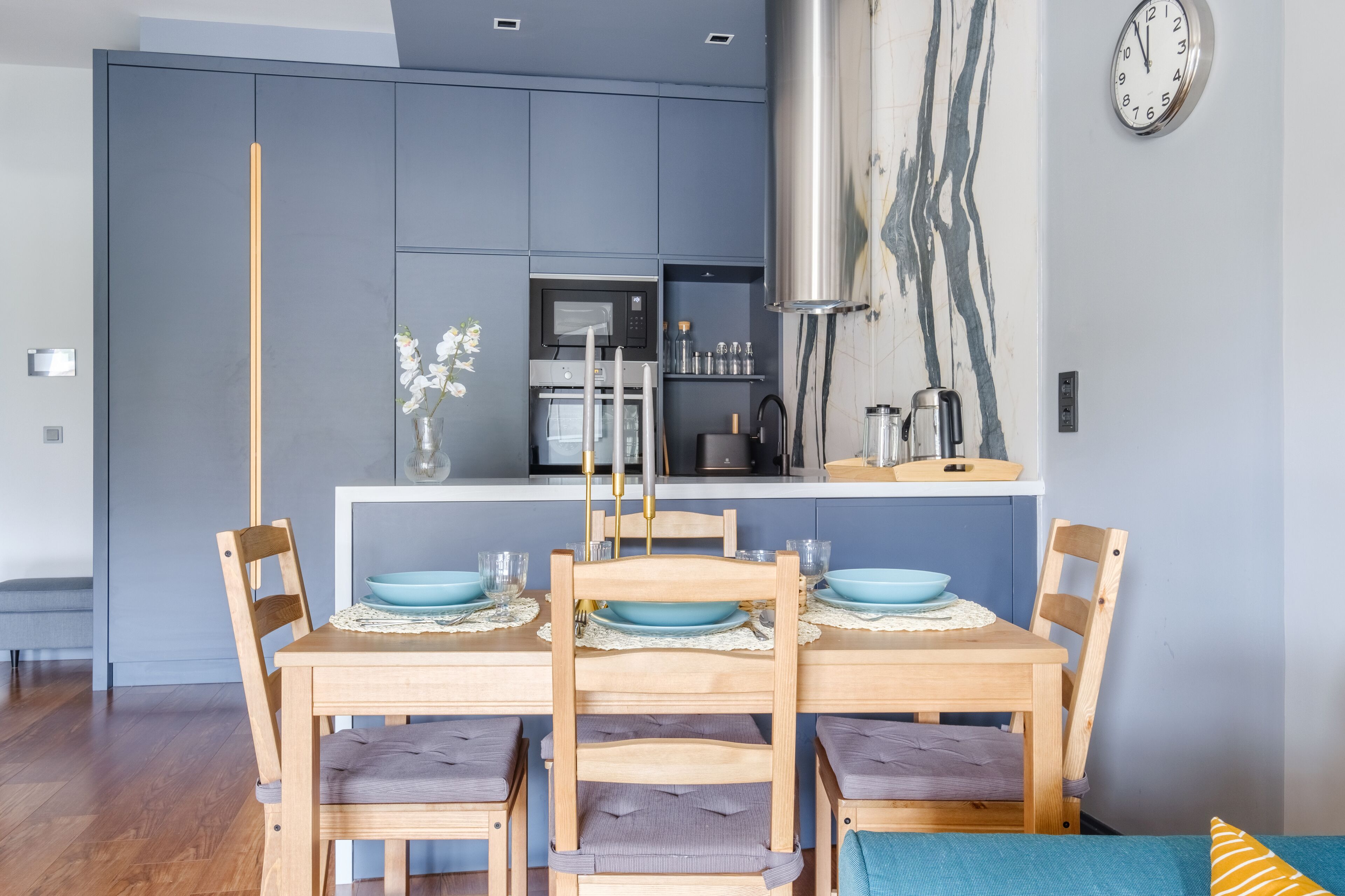 A dining area with a wooden table set for four, a kitchen in the background, and a clock on the wall.