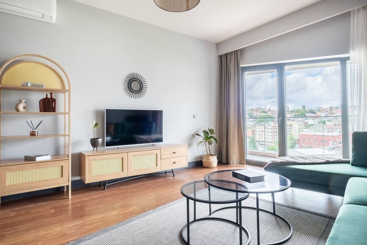 A modern living room with a TV, wooden furniture, a glass coffee table, and a large window with a city view.