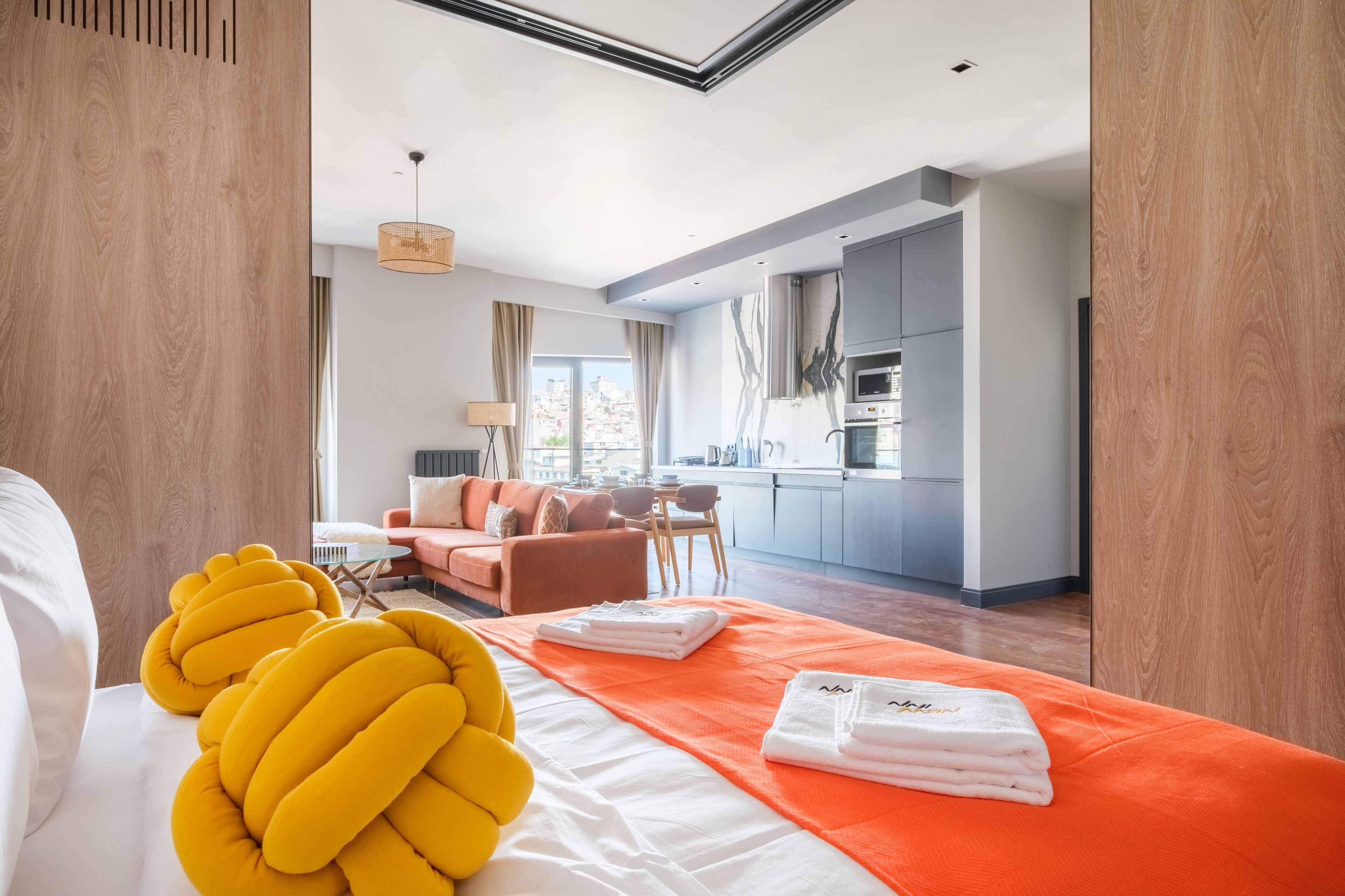 A modern hotel room with a large bed, a sofa, a dining area, and a kitchen in the background.