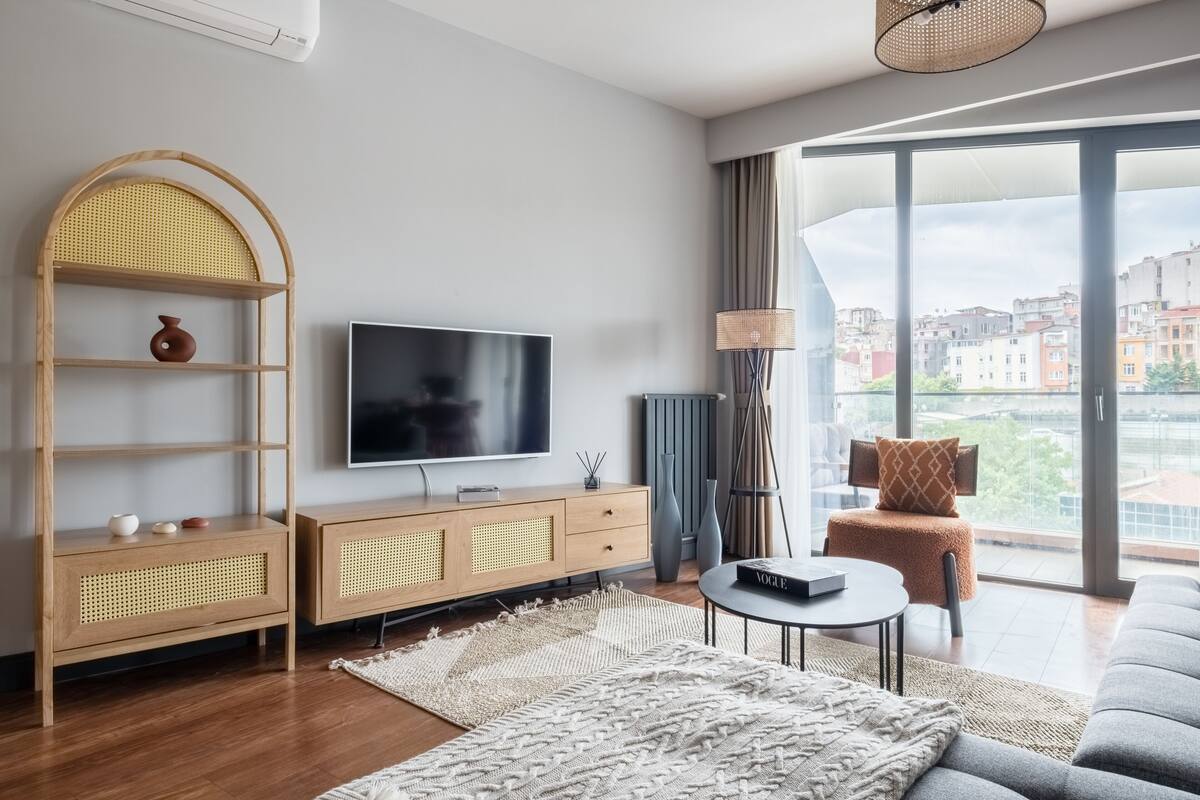 A modern living room with a large TV, wooden furniture, a sofa, and a view of buildings through large windows.