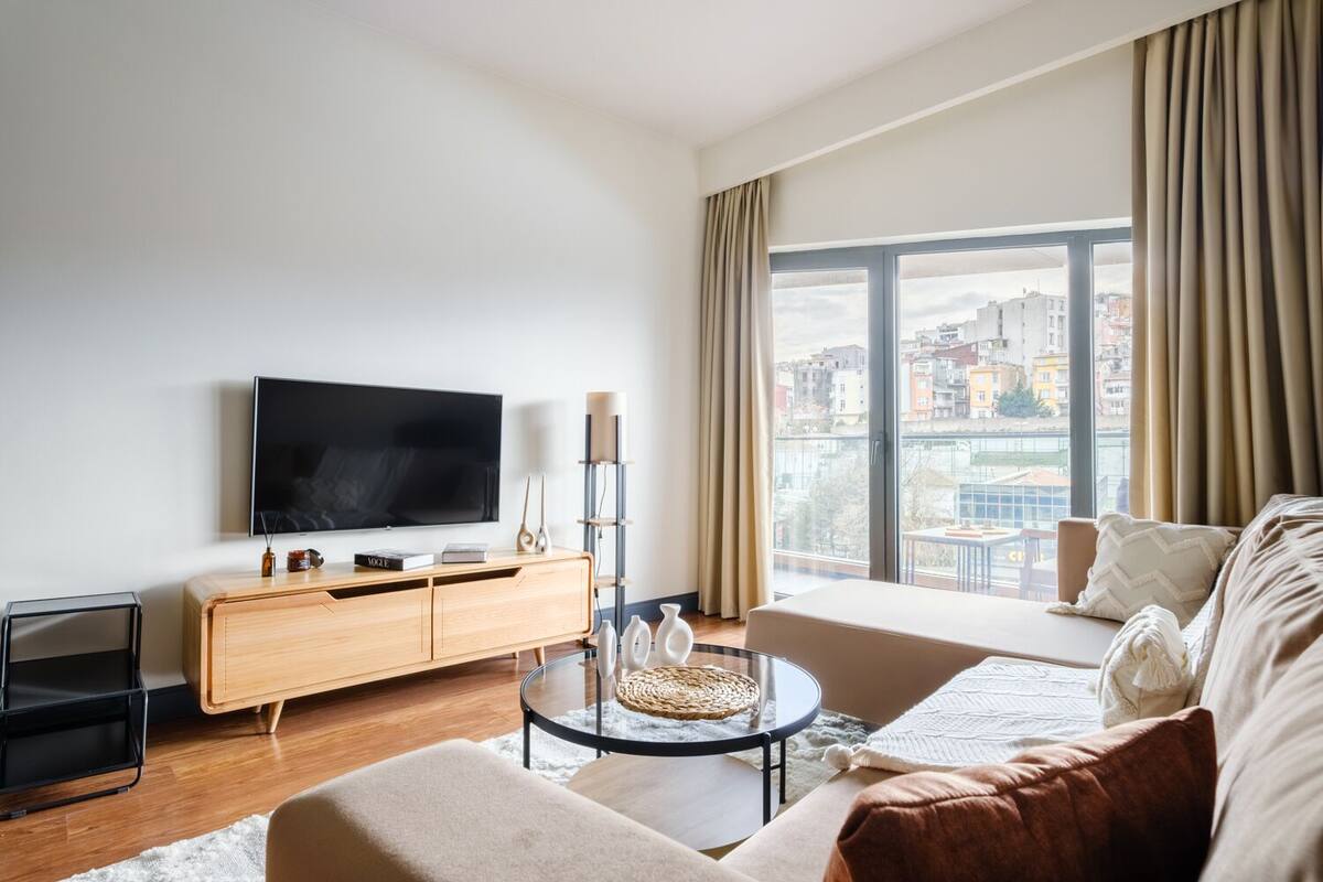 A modern living room with a flat-screen TV, a wooden cabinet, a glass coffee table, and a sofa with a view of the city through large windows.