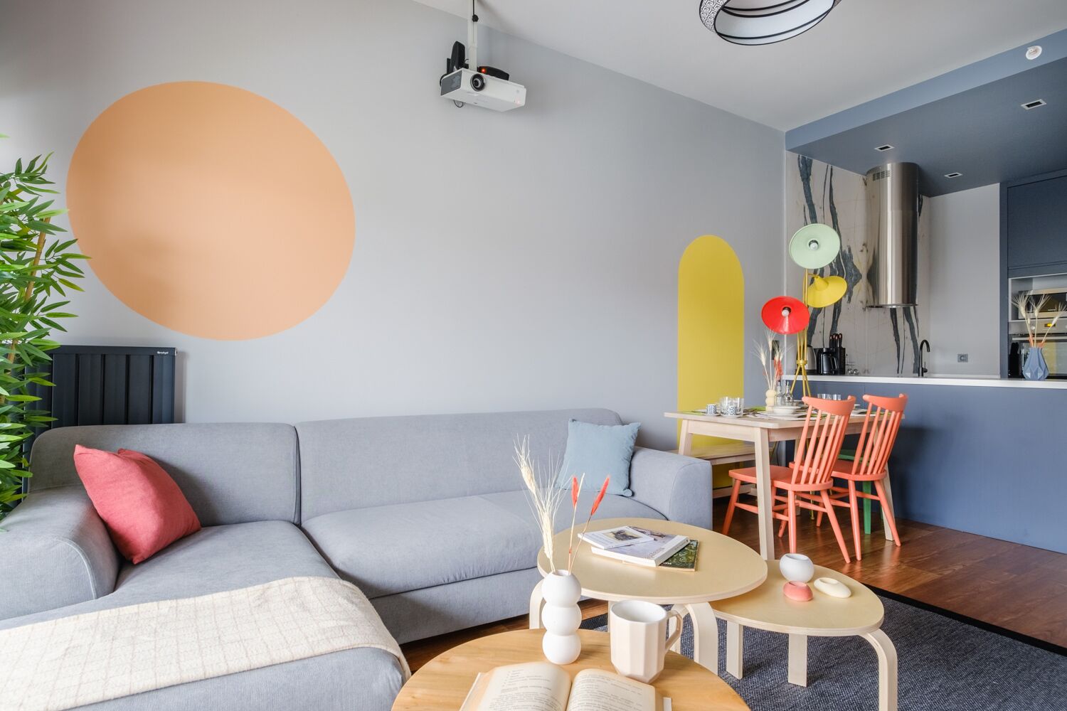 A modern living room with a grey sofa, a coffee table with books and a vase, a dining area with a small table and chairs, and a kitchen area with a stove and cabinets.