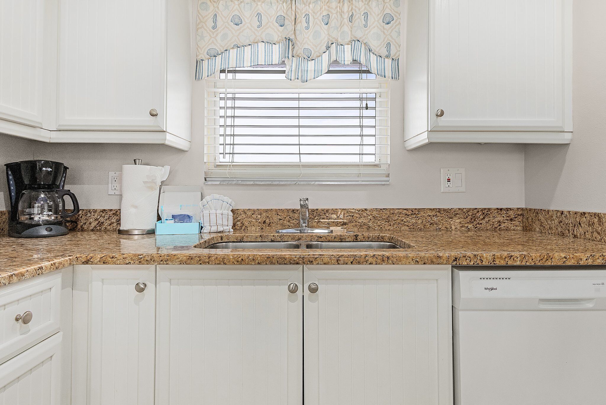 Una cocina con armarios blancos, encimera de granito, cafetera, lavabo y ventana con persianas.