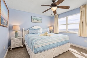 A bedroom with a bed, nightstands, a ceiling fan, a window with curtains, and a beach painting on the wall.