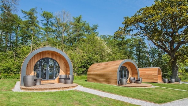 Dos cabañas tipo cápsula de madera con ventanas amplias y zonas de estar, rodeadas por un césped cuidado y árboles.