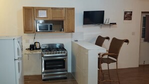 A compact kitchen with a microwave, stove, and refrigerator. There is a TV mounted on the wall and a small shelf above it. Two wicker chairs are placed near the kitchen counter.