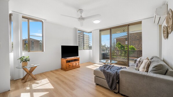 A modern living room with a flat-screen TV, a glass coffee table, a grey sofa, and a ceiling fan.