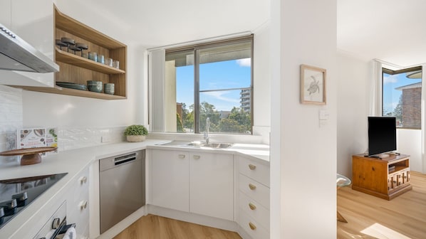 A modern kitchen with white cabinetry, a built-in dishwasher, a window with a view, and a wooden cabinet with kitchenware.