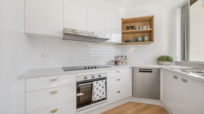 A modern kitchen with white cabinetry, a built-in oven, a dishwasher, and a wooden shelf above the countertop.