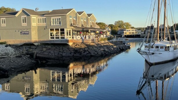 A waterfront property with a large house featuring multiple windows and a balcony, a dock with a sailboat, and a calm body of water reflecting the buildings. - Historic Kennebunkport home only 0.3 miles to Dock Square (Kennebunkport)