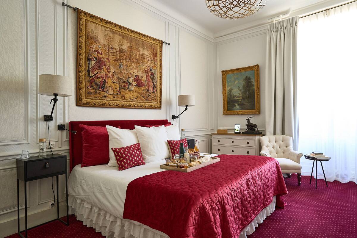 Ein Hotelzimmer mit einem großen Bett, zwei Nachttischen, einer roten und weißen Bettdecke, einem großen Gemälde an der Wand, einem Sessel, einem kleinen Tisch und einem Fenster mit Vorhängen.