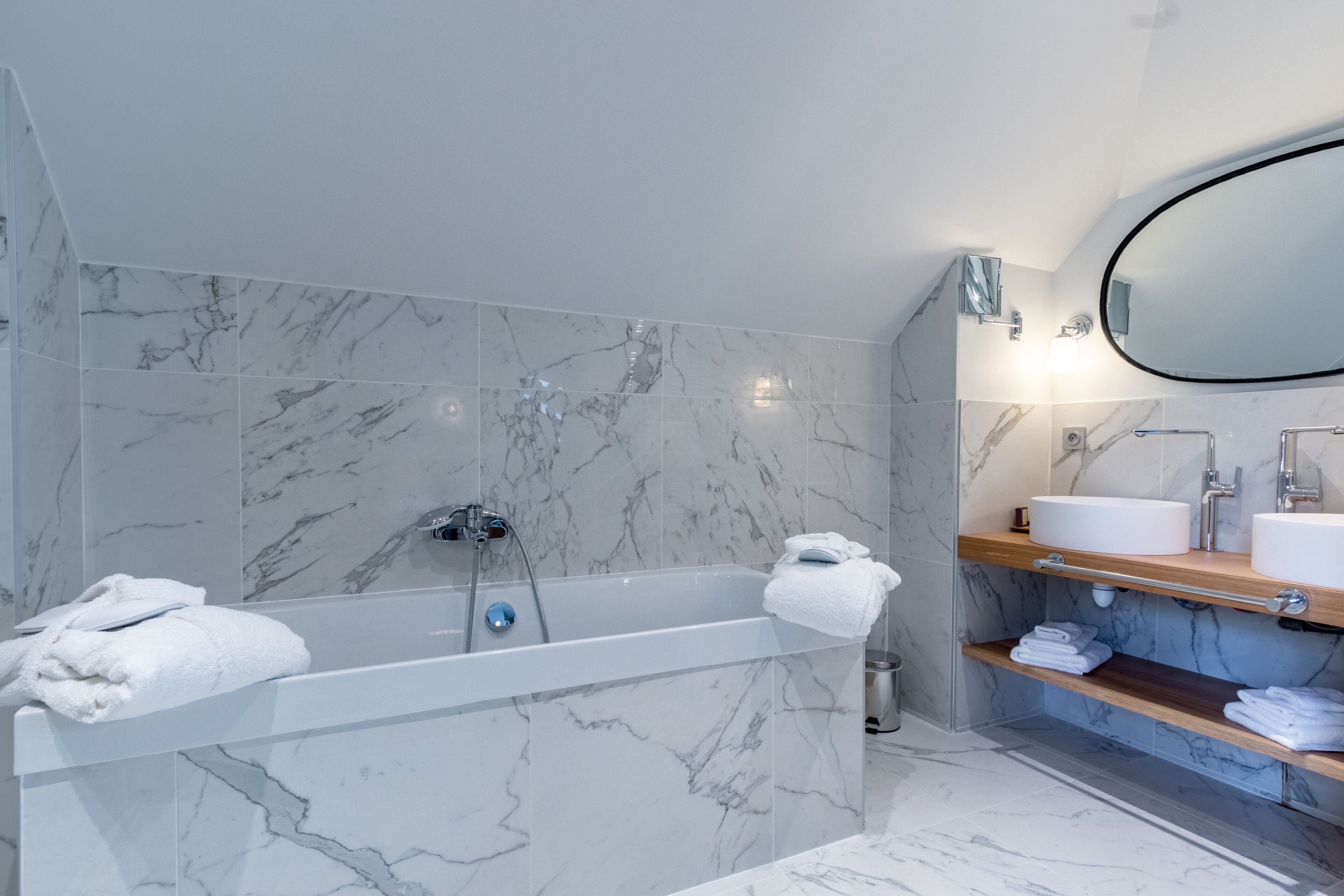 A modern bathroom with a marble-tiled wall, a freestanding bathtub, a wooden vanity with two sinks, and a large mirror.