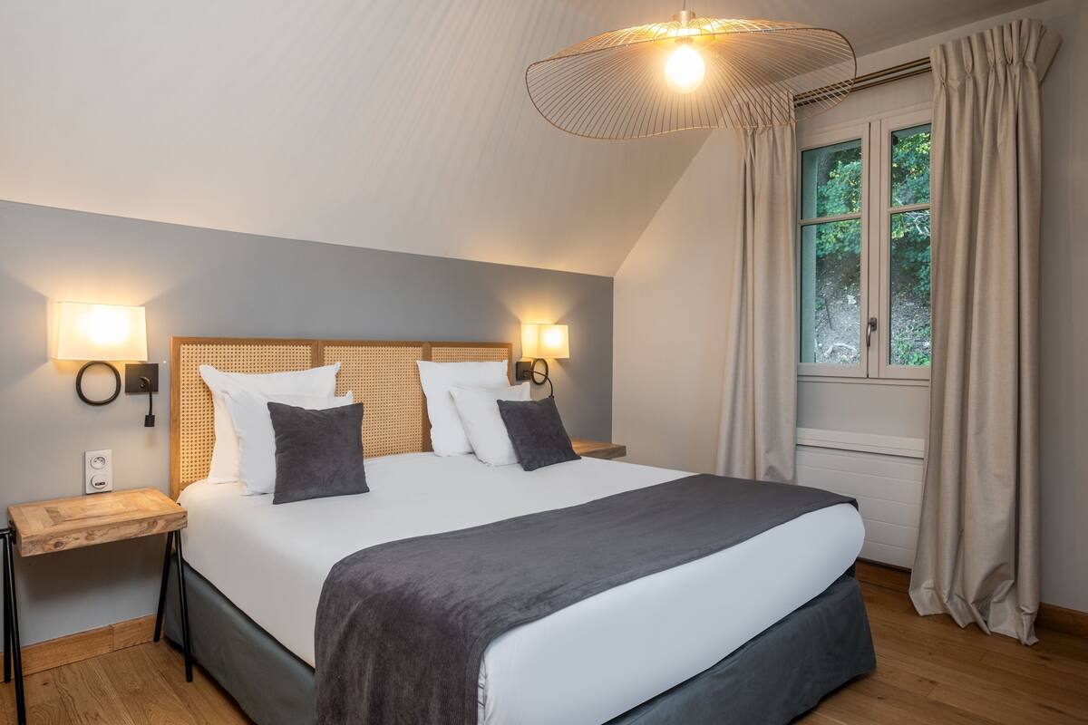 Ein ordentlich bezogenes Bett mit grauer Decke, zwei Nachttische mit Lampen, ein Holz-Kopfteil und ein Fenster mit Vorhängen.