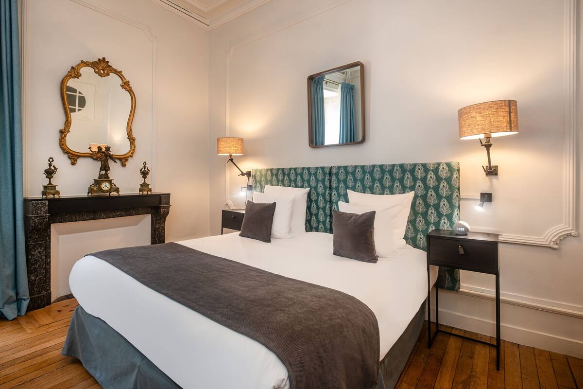 Ein Hotelzimmer mit einem großen Bett, zwei Nachttischen, einem Spiegel, einem Kamin und einer Uhr.