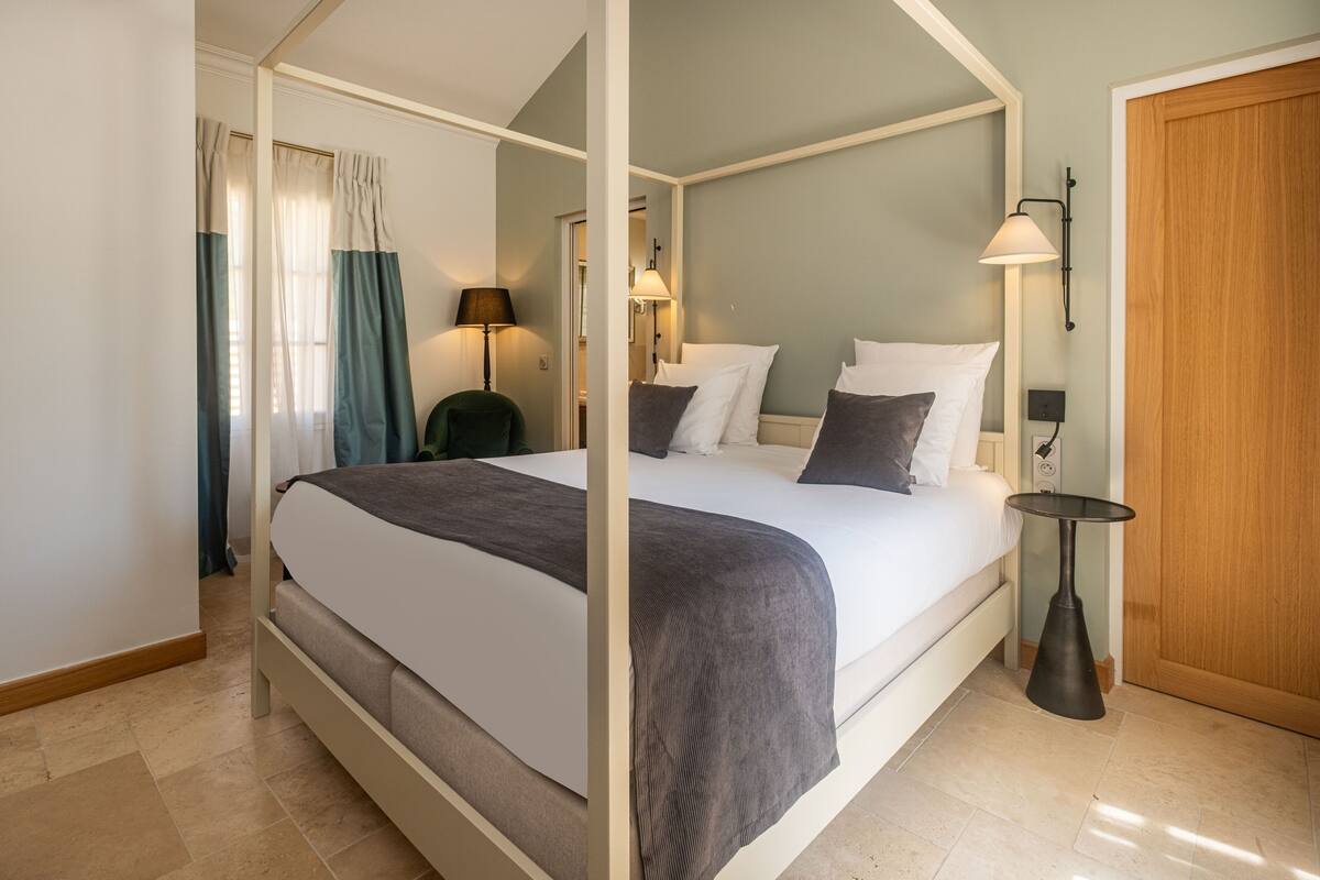Ein Himmelbett mit grauer Bettdecke, ein Nachttisch mit Lampe und eine Holztür.