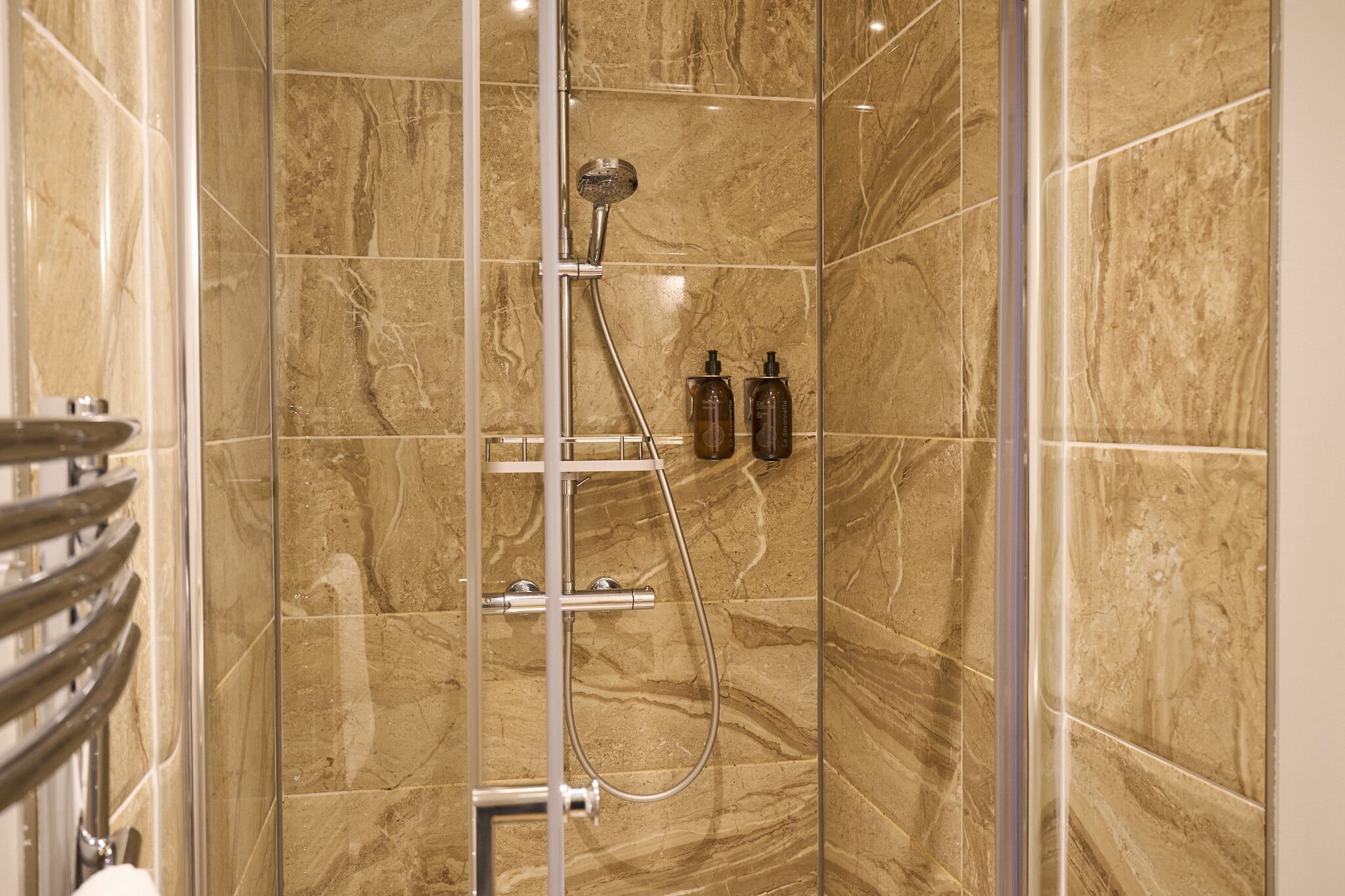 A shower with a glass door, marble tiles, and two bottles of shampoo.